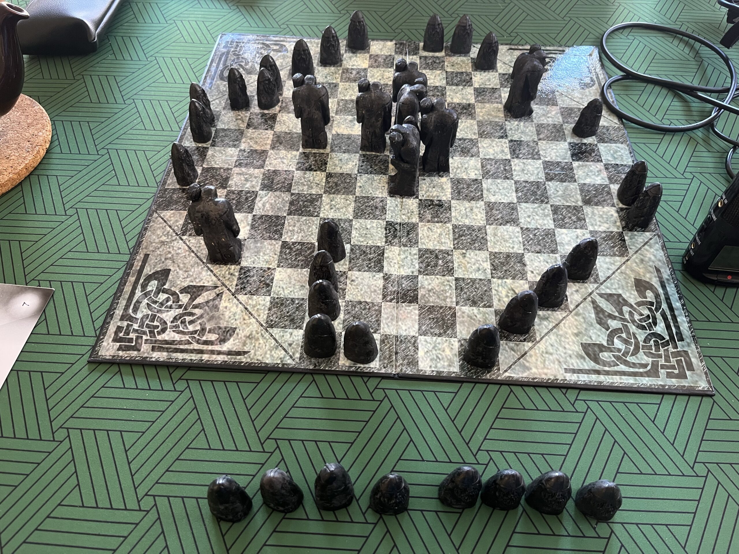 A square board with an octagonal arrangement of black and white squares sits on a green patterned matt on a wooden table. On the board are dark-coloured playing pieces, some large, humanoid shaped ones holding up clubs, and some smaller, stylised bullet-shaped pieces with bearded faces. There are many of the smaller pieces on squares around the outside edge, while the larger ones are all near the centre. Eight of the smaller pieces are off the board, on the matt closer to the camera.