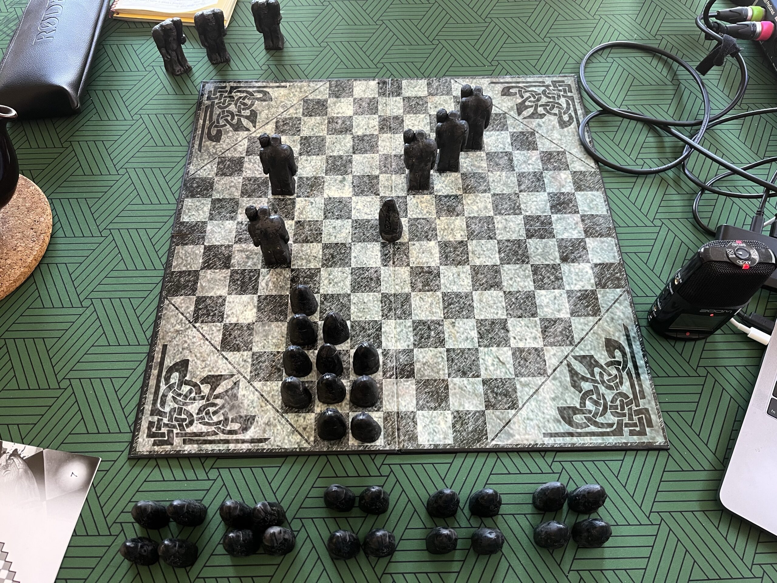 A square board with an octagonal arrangement of black and white squares sits on a green patterned matt on a wooden table. On the board are dark-coloured playing pieces, some large, humanoid shaped ones holding up clubs, and some smaller, stylised bullet-shaped pieces with bearded faces. The smaller pieces are in a group on the bottom edge of the board, while the larger ones are on the left and top right of the board. Eight of the smaller pieces are off the board, on the matt closer to the camera. About two-thirds of the smaller pieces are off the board at the bottom, close to the camera, while three of the larger pieces are off the board at the top.