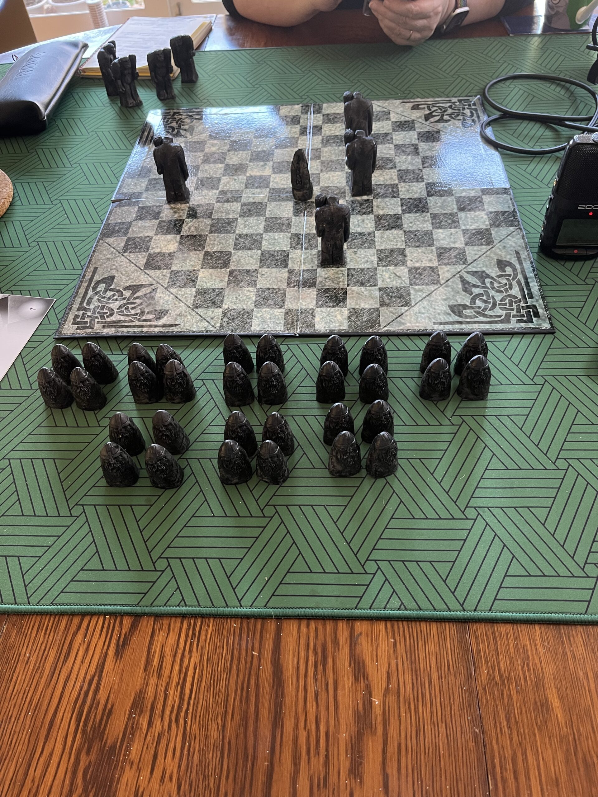 A square board with an octagonal arrangement of black and white squares sits on a green patterned matt on a wooden table. On the board are four large, dark-coloured playing pieces, humanoid-shaped and holding clubs. There's also a pointy piece in the centre of the board. Four more of these pieces are off the board at the top, while 32 smaller pieces with points are clustered in groups of four off the board at the bottom, near the camera.