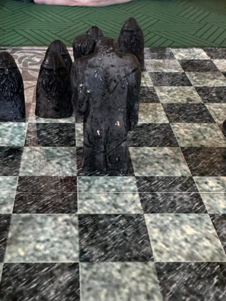 A large black playing piece of roughly humanoid shape sits on a black square on a board that resembles a chess board, with some other smaller pieces behind it. The humanoid one faces away from the camera and you can see its butt. It's a troll's butt.