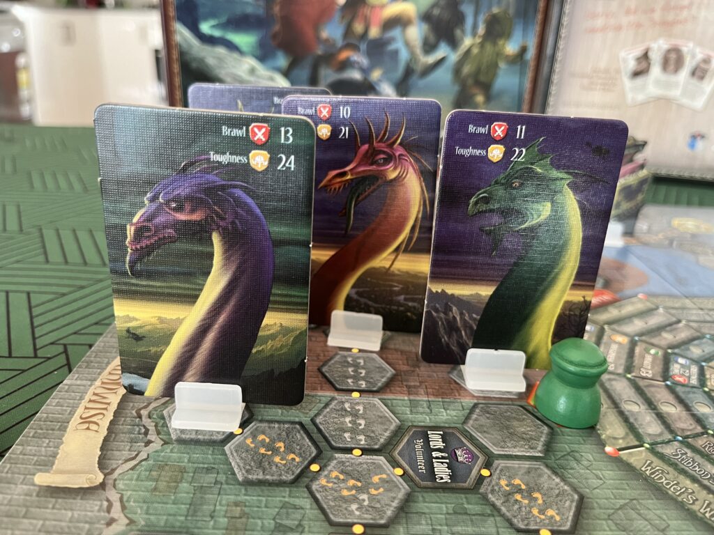 A photo of the four cardboard standees for the dragons in the game, on the board next to a player’s pawn. Three of them are clearly visible: the illustrations are of the head and long neck of a purple, red and green dragon.