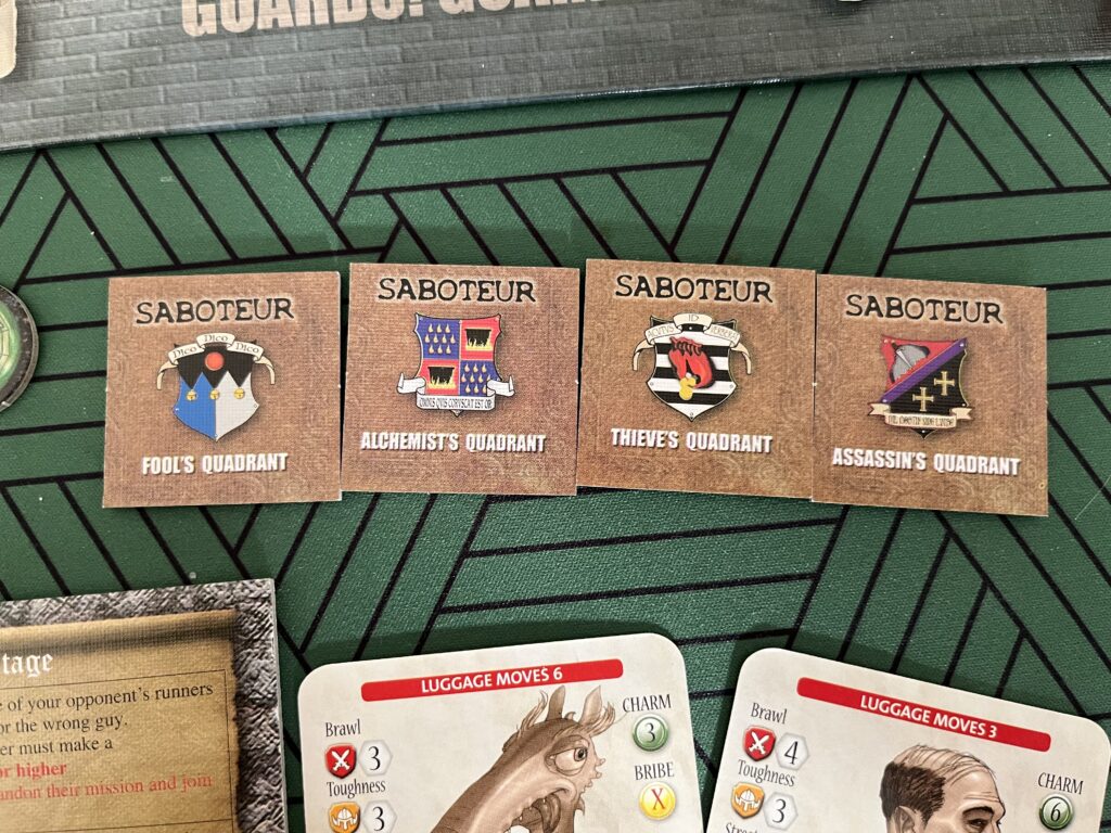 A photo of the four Saboteur markers face up, showing the crests of the four different guilds in the game, and labelled “SABOTEUR” above and the name of the quadrant: the Fool’s, Alchemist’s, Thieve’s and Assassin’s Quadrants. Some other game components are visible at the edges of the photo.