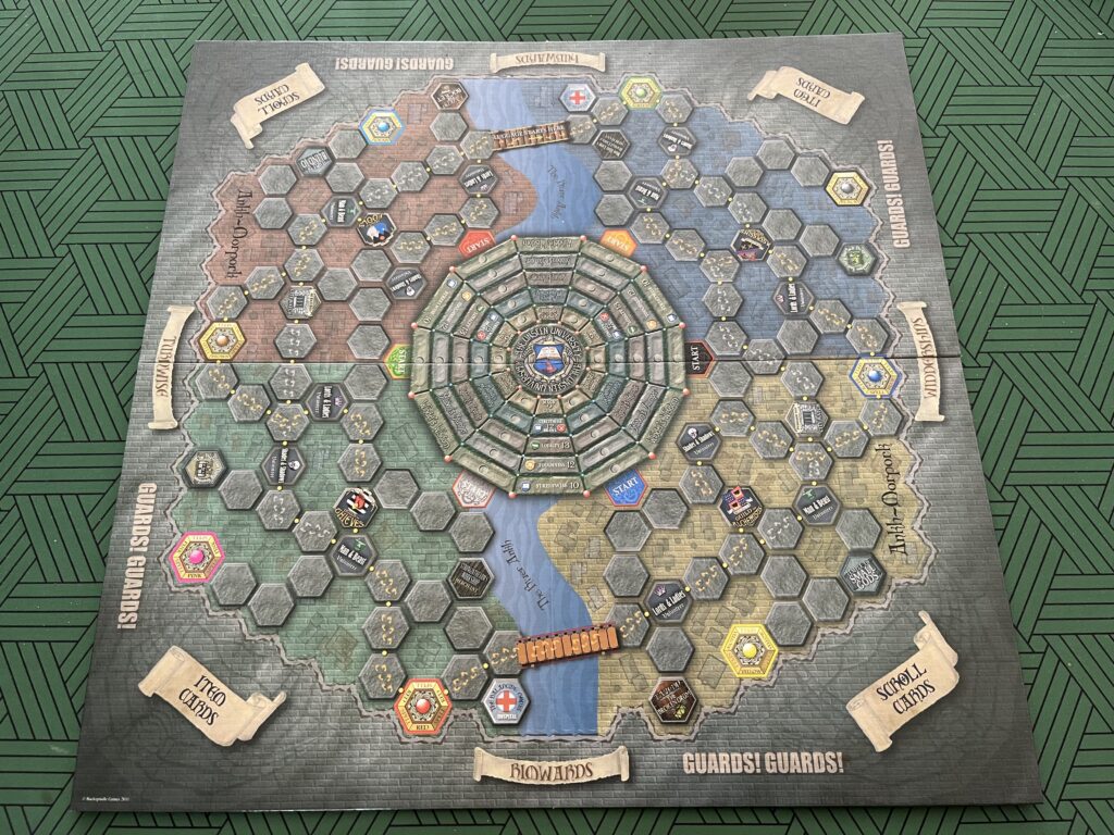 A photo of the Guards! Guards! board from above, showing the twelve-sided central section representing Unseen University, the River Ankh dividing the board into two halves, and the pattern of hexagonal spaces representing the city streets and various special locations. Around the outside of the board are labels showing where piles of item and spell scroll cards should go, as well as the cardinal directions of the Disc.