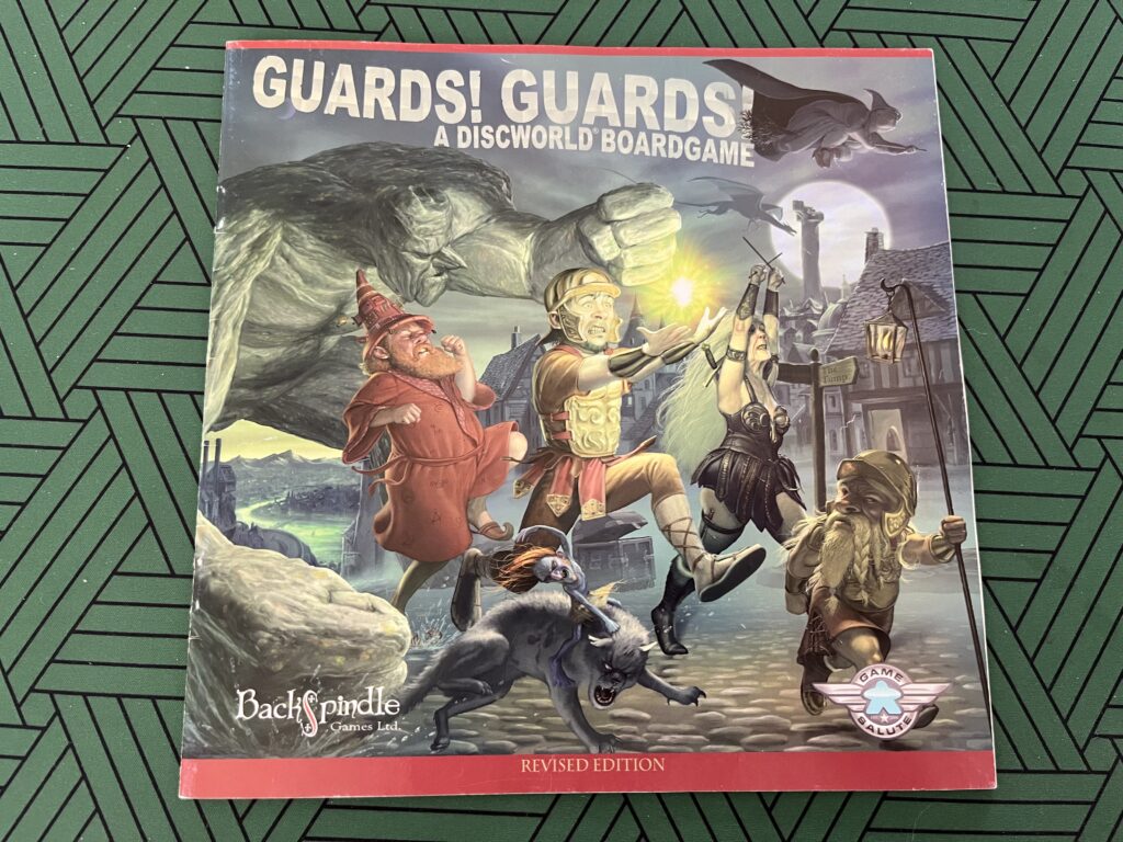 The Guards! Guards! rulebook. The illustration on the cover shows a member of the City Watch in breastplate, leather skirt and helmet running while carrying a glowing mote of light which hovers just above his hands. He is surrounded by various other characters running with him through the streets of Ankh-Morpork at night, including a red-bearded wizard in a red robe, a Feegle riding a cat, a dwarf, an older woman in black leather armour and a huge rocky troll. A classic witch silhouette flies through the sky in the background above a full moon.