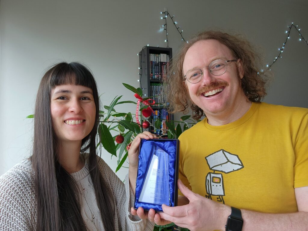 A photo of a two people smiling at the camera: a woman with long dark straight hair in a light grey knitted jumper, and a man with mid-length long curly red hair, moustache and glasses wearing a yellow T-shirt with an illustration of sad robot on it. They are holding an open box linked in blue material containing a trapezoid-shaped glass trophy with writing etched into it. The writing is not legible. In the background are some Christmas lights and an Australian native tree decorated with Christmas baubles.