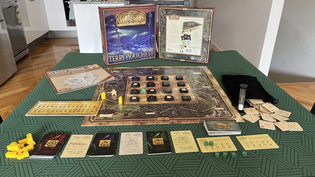 A photograph of the Clacks board game components laid out on a small table in front of the box. These include the board; square wooden lamp tokens; small wooden “meeples” - humanoid playing pieces - for one player and for the Post Office; a cardboard “Deep Dwarf” token; wooden Clacks tower pieces; the game manual; the large Clacks code reference card; a cloth bag filled with square jacquards; an egg timer; and the cards for Clacks message words, fault and incident reports, and operator’s logs.